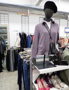 Kuvassa on mallinukke. Taustalla on kierrätysmyymälän vaaterekkejä.